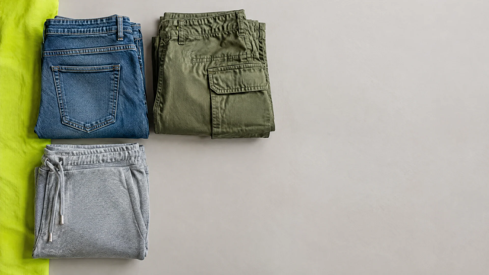 Folded blue jeans, olive cargo pants and heather gray joggers laid out in a grid next to a lime green strip